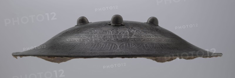 Shield sipar, Persian Iran, Qajar dynasty 1785–1925, 18th/19th century. Metal weapon, diameter 47.5 cm, height 9.0 cm. Decoration: field with hunting scenes horses, archers, belts ornamented with plant tendrils and inscriptions stylized in Kufi script. Function: militaria/hunting., A shallow, rounded metal object with a domed top and a flared rim sits against a plain light gray background; the metal surface is dark gray with engraved decorative patterns and a few raised rounded knobs on the top, and a soft, frayed material in beige and light brown is attached beneath the rim.