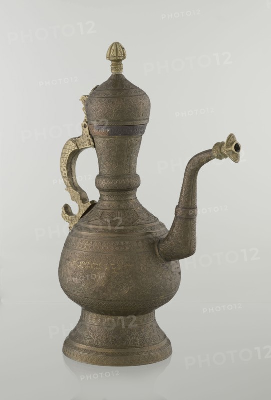 Jug aftabe, aftabe, geometric and plant ornament, dragons, zoomorphism; dense plant and geometric decoration; Persian, Qajar dynasty 1785–1925, 19th century; Central Asia, / Iran ; vessel, height 34.0 cm, width 22.7 cm, diameter 13.3 cm., An ornate metal ewer with a bulbous body, a flared foot, a long curved spout, a tall neck with a domed lid topped by a finial, and a hinged decorative handle; the surface is covered in dense engraved patterns and textured motifs, and the handle and finial show a brighter metallic tone while the main body and spout appear darker with a patinated finish in shades of bronze, gold, and brown against a pale gray background.