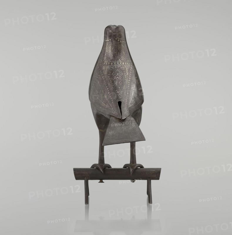 Falcon on a branch, Persian, 18th century, Iran; figurine sculpture depicting a falcon with feathers, inscriptions/calligraphy; height 28.5 cm, width 27.5 cm, depth 14.5 cm, weight 1.25 kg., A dark metallic bird-like sculpture stands on a narrow horizontal perch with a few supporting legs; the sculpture has an elongated body with a decorative patterned surface, a triangular piece extending from its lower front, slender legs and clawed feet gripping the perch, and a smooth rounded head. The visible colors are dark gray, black, and hints of brown, all set against a light gray background.