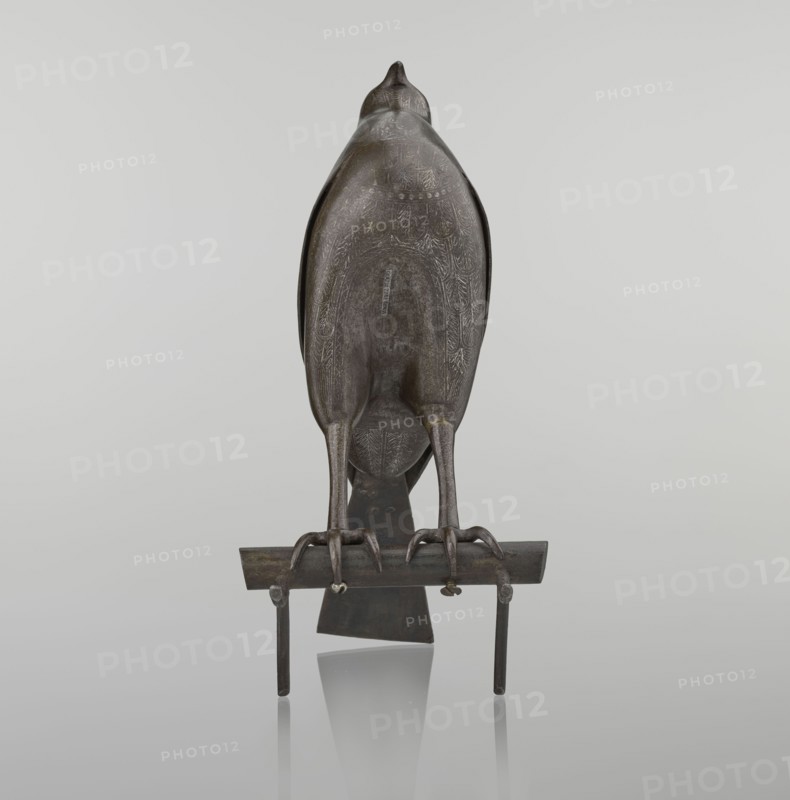 Falcon on a branch, Persian, 18th century, Iran; sculpture/figurine of a bird falcon with feathers, inscriptions/calligraphy; figurine; height 28.5 cm, width 27.5 cm, depth 14.5 cm, weight 1.25 kg., A sculpted bird figure viewed from behind, perched on a horizontal bar supported by vertical posts, with carved detailing on its back and wings; the bird and stand have a dark metallic brown-gray surface with subtle lighter highlights and a few areas of patina that appear greenish and yellowish, set against a smooth pale gray background with a soft reflection beneath the stand.