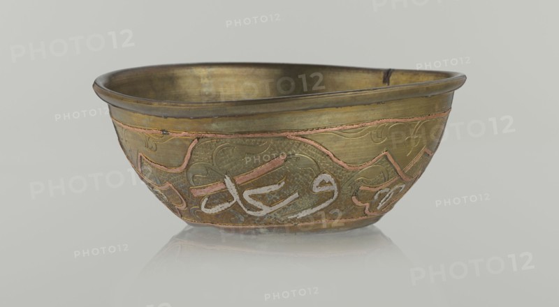 Bowl, Islamic art, Arabic culture, arabesque ornament, calligraphy, Arabic inscriptions, geometric motifs, 19th century, Middle East, possibly Syria, vessel, height 4.9 cm, diameter 13.3 cm, A shallow metal bowl sits against a pale gray background, shown in a frontal view with its interior visible; the bowl has a warm brass color with areas of darker patina and a few copper-colored inlaid lines forming decorative shapes, and parts of its surface show a slightly textured, hammered appearance; there are traces of a pale grayish-white material in curved strokes on the exterior decoration, and the rim is slightly darker with subtle wear; the bowl rests on a reflective surface that creates a faint mirrored reflection beneath it.