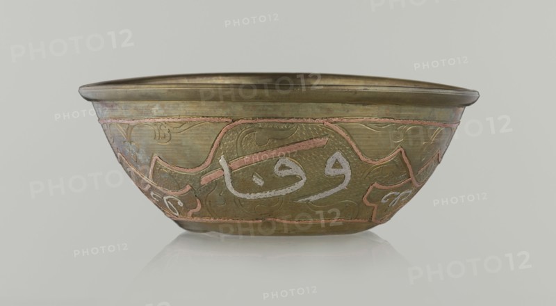 Bowl, gift, Islamic art, Arabic culture, arabesque ornament, calligraphy, Arabic inscriptions, geometric motifs, 19th century, Middle East, possibly Syria, vessel, height 4.9 cm, diameter 13.3 cm, A shallow metal bowl with a rounded rim sits against a pale gray background; the bowl shows a predominantly brass-colored surface with areas of darker and lighter patina, and it is decorated with a band of raised curving designs in a pinkish tone and a few pale silvery accents; the body of the bowl has textured hammered areas and smooth sections, with ornamental flowing motifs in subtle engraved lines; the rim and base edges are slightly darker brass, and the overall color palette includes brass, pinkish copper, pale silver, and muted greenish-brown tones.