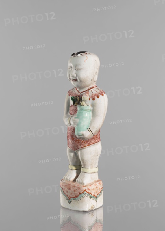 One of the Hehe Er Xian brothers holding a vase with lotus buds;, Chinese, Qing dynasty, Kangxi period 1662–1722; colored porcelain sculpture; subject: boys/children and lotuses; height 28.0 cm; weight 0.75 kg; dated 1662–1722; China., A ceramic figurine of a standing child on a decorated pedestal, shown against a plain gray background. The child has a pale off-white complexion with subtle shading, short dark hair painted on top of the head, and a smiling facial expression with closed eyes. The figurine wears a red ornamental collar with petal-like shapes and a red patterned loincloth, and pale yellow anklets around the lower legs. The child holds a glossy light green vase with a few green leaves and a darker element at the top. The pedestal is mainly off-white with a pinkish-red patterned top and scalloped green and red decorative bands along the sides. Colors visible include off-white, gray, red, pinkish-red, green, light green, dark green, yellow, and black.