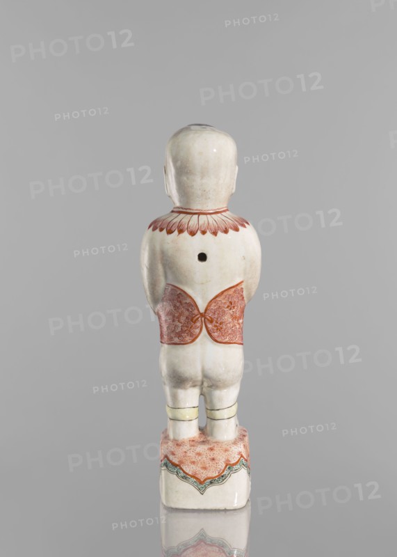 One of the Hehe Er Xian brothers holding a vase with lotus buds, Chinese, Qing dynasty Kangxi, 1662–1722, colored porcelain sculpture of boys/children with lotuses, height 28.0 cm, weight 0.75 kg, dated 1662–1722., A small pale figurine viewed from behind standing on a rectangular base, with a smooth rounded head and visible ears, a short neck rimmed by petal-like decoration in reddish tones, a cylindrical torso with a dark circular hole on the upper back, a decorative reddish patterned band wrapped around the lower torso, short legs with pale yellow bands near the knees, and a base decorated with a mottled reddish surface and scalloped green and reddish edging; visible colors include pale off-white, reddish, yellow, green, and hints of gray.