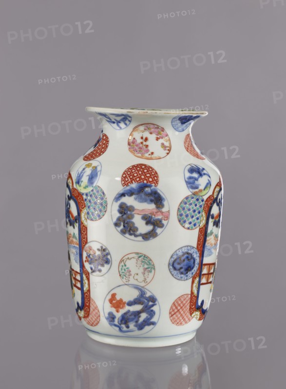 Vase, Tashiro maker/trading company, Japanese, colored porcelain, ceramics, Meiji period 1868–1912, 2nd half of 19th century. Hizen, Arita, Japan. Height 28.5 cm; diameter 19.0 cm. Decoration: figurative scenes with landscape background, peonies, camellias, wisteria, fish, pine trees, asanoha pattern. Provenance unknown., A white ceramic vase decorated with a pattern of round medallions and vertical panel shapes, featuring painted scenes and motifs in blue, red, green, pink, and gold tones, with a flared rim and a gently tapered body standing on a short base, set against a plain gray background.
