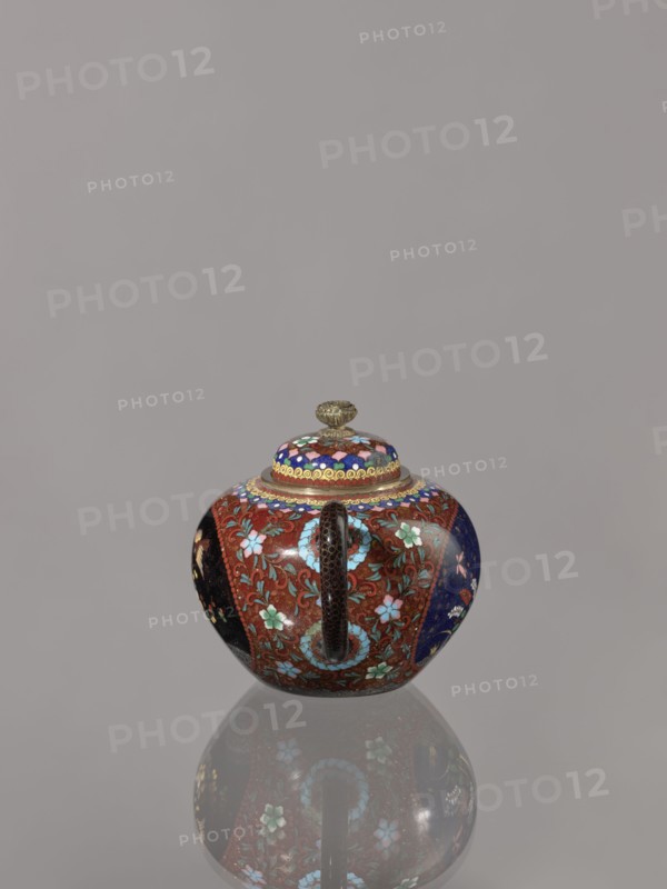 Teapot kyûsu with lid, butterflies, plant motifs, Japanese culture, birds, plum trees, Meiji 1868–1912, lids, cloisonné enamel. The lid is topped with a stylised lotus flower. Decoration: two reserves on the belly — a plum tree and a bird; plants and butterflies; the rest of the background and the cover — a floral tendril. Date ca. 1906–1908 Meiji 39–41. Japan, Kyoto. Vessel. Height 10.0 cm, diameter 10.0 cm., A small rounded lidded vessel with a central vertical handle sits on a reflective surface, showing a mirrored reflection beneath it. The vessel is richly decorated with floral and scrolling patterns in colors including red, blue, turquoise, green, pink, black, gold, and purple. The lid has concentric decorative bands and a small ornate gold-colored finial. The body displays panels of different background colors with flower motifs and curving vine designs, and the handle has a dark textured appearance. The background is a plain neutral gray.