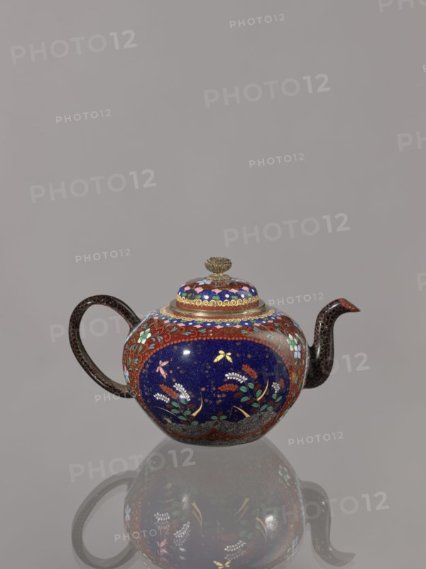 Teapot kyûsu with lid;, Cloisonné enamel; ca. 1906–1908 Meiji 39–41, Kyoto, Japan. Height 10.0 cm, diameter 10.0 cm. The lid is topped with a stylised lotus flower. Decoration: two reserves on the belly showing a plum tree and a bird, with plants and butterflies; the remainder of the background and the cover decorated with a floral tendril. Subjects: birds, plum trees, plant motifs, butterflies., A rounded teapot with a curved handle and spout sits against a gray background, showing a reflective surface beneath. The teapot features a dominant deep blue central band decorated with floral motifs in green, white, pink, yellow, and red, surrounded by reddish-brown borders with floral and leaf patterns. The lid has concentric decorative rings in blue, red, and gold topped by a small gold-colored finial. The handle and spout are dark brown with a subtle dotted texture. The overall colors visible include blue, red, brown, gold, green, white, pink, yellow, and gray.