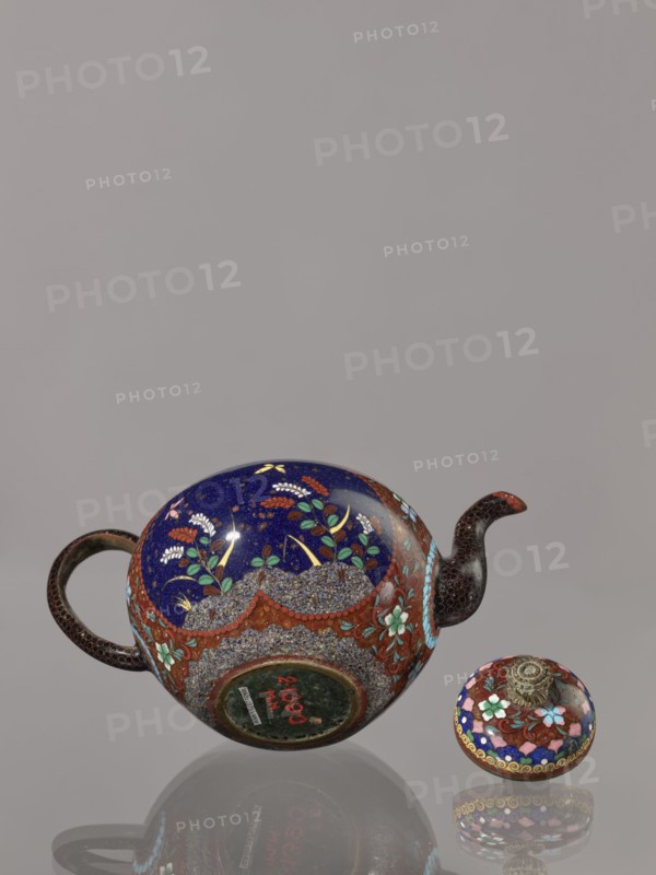 Teapot kyûsu with lid;, Cloisonné enamel teapot, ca. 1906–1908 Meiji 39–41, Kyoto, Japan. Height 10.0 cm, diameter 10.0 cm. The lid is topped with a stylised lotus flower. Decoration: two reserves on the belly—a plum tree and a bird—plus plants and butterflies; the rest of the background and the cover decorated with a floral tendril. Subjects: birds, plum trees, butterflies, plant motifs., A rounded teapot and a separate lid rest on a reflective surface against a plain grey background. The teapot has a deep blue upper surface with floral motifs in green, white, yellow, and red, and a band of textured, pebble-like patterning in grey and brown around its midsection accented with red and turquoise floral decorations. The handle and spout are dark brown with a subtle scale-like or dotted texture, and the spout tip shows a small area of red. The separate lid echoes the teapot’s colors with blue, red, turquoise, white, and gold decorative patterns and a central metallic, sculpted finial in a...