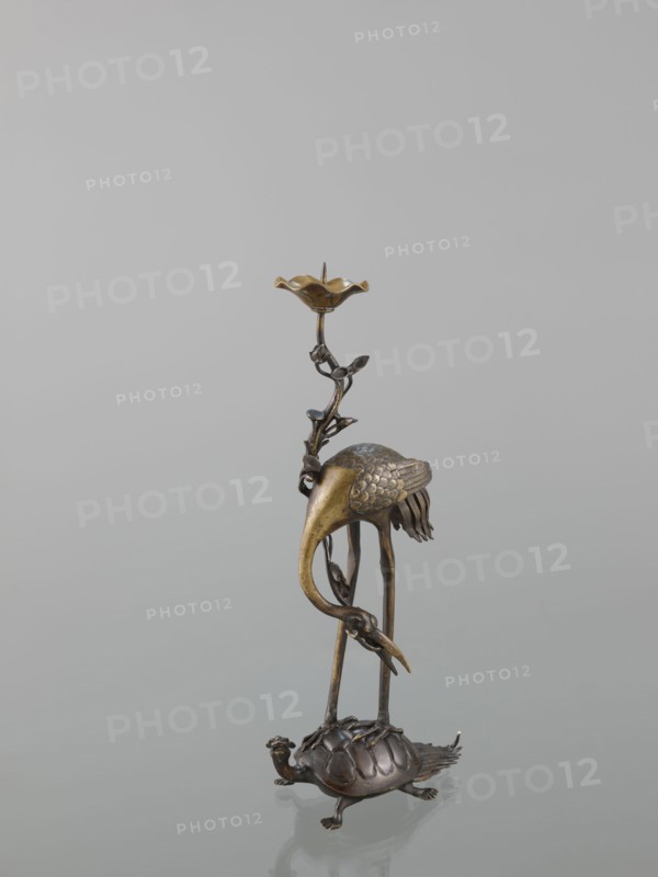 Candlestick shokudai, ; materials/features: lotuses, turtles, cranes iconography, animals; style/period: Meiji 1868–1912, 2nd half of the 19th century, Japan; function: candlestick; height: 37.0 cm., A metallic sculpture showing a tall bird with a long curved beak and textured wings standing on thin legs that rest on the back of a turtle; a decorative stem with small leaf-like elements rises beside the bird and ends in a shallow flower-shaped dish at the top. The sculpture has a mix of bronze, gold, and dark brown tones against a pale gray background.