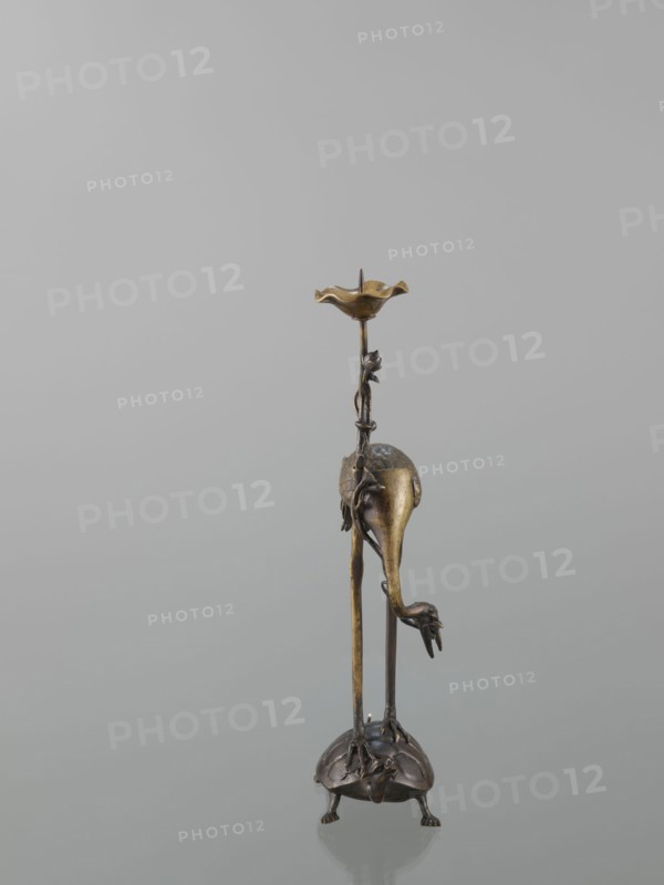 Candlestick shokudai, ; materials/features: lotuses, turtles, cranes iconography, animals; culture: Japanese, Meiji 1868–1912, 2nd half of the 19th century, Japan; height 37.0 cm., A slender metal sculpture with a muted gold and dark bronze finish stands on a rounded base that resembles a turtle form with textured shell-like details and small feet; two long, thin vertical supports rise from the base, one of which curves into a drooping elongated shape that ends in a clawed foot, while a vine-like element wraps partway up the supports and carries a small decorative flower near the top; the uppermost section features a shallow petal-shaped dish with a scalloped edge in a warm gold tone; the background is a smooth, neutral gray and the surface beneath the sculpture shows a soft reflection.
