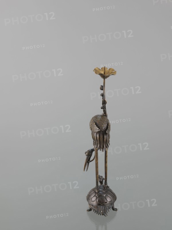 Candlestick shokudai, lotuses, turtles, cranes iconography, Japanese art, Japanese culture, Meiji 1868–1912, animals, 2nd half of the 19th century, Japan, candlestick, height 37.0 cm, A slender metallic sculpture stands against a pale gray background, showing a vertical form with a base resembling a rounded shell or dome with textured segments and fringe-like elements at its lower edge; from this base rise long thin supports leading up to a bell-shaped cluster of layered, petal-like pieces and dangling fringe, with a small bird-like figure perched on one of the supports; higher up a twisted vine-like stem with tiny leaf shapes ascends to a flared, shallow cup at the top. The visible colors are bronze, gold, dark brown, and muted gray.