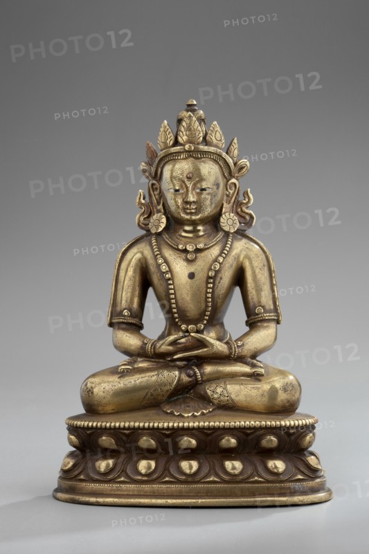 Amitayus, Himalayan/Tibetan Buddhist and Chinese/Mongolian cultural influences, lotus throne iconography; Buddha Amitayus seated on a lotus or vajra throne, holding a vase of long life; 18th–19th century; figurine sculpture and cult object; height 11 cm, width 7.8 cm, depth 5.5 cm., A seated metallic statue with a golden and slightly darker patina shows a figure in a cross-legged pose on an ornate rounded pedestal; the figure wears an elaborate crown with petal-like projections, large circular earrings, and layered beaded necklaces, and its hands rest together at the lap. The surface displays a mix of bright gold and darker brownish-gold tones with some worn areas, and the pedestal features repeating raised teardrop shapes and beaded trim in similar golden and brown hues. The background is a smooth neutral gray.