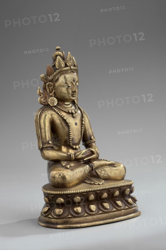 Amitayus, Tibetan Buddhist/Himalayan and Chinese art influences, lotus throne, Buddha Amitayus holding a vase of long life, seated lotus or vajra posture, 18th–19th century, possibly China or Mongolia, figurine sculpture and cult object; height 11 cm, width 7.8 cm, depth 5.5 cm., A seated metallic statue on an ornate base, shown against a plain gray background, with a golden-bronze surface that shows variations of darker tarnish and lighter highlights; the figure wears a decorative crown with petal-like elements and a small pointed finial, has stylized hair and large circular earrings, and displays a calm facial expression with closed or downcast eyes and a raised mark on the forehead. The torso is adorned with layered beaded necklaces and armlets, and the arms rest in the lap with hands arranged in a meditative gesture. The clothing details include patterned bands around the legs and a draped element over one shoulder. The rounded base beneath the figure features a repeating petal mot...