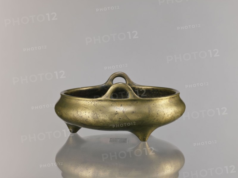 Li-type incense burner, Chinese, bronze ritual vessel, Ming dynasty Xuande period, 1426–1435, China; incense burner; height 10.0 cm, diameter 23.5 cm, A small metallic vessel with a rounded, bowl-like body and a dull gold surface sits on a reflective surface, showing a faint mirror image beneath; the vessel has a curved rim, two arching loop handles that rise from opposite sides and meet near the center, and it rests on three short tapered feet; the metal shows mottled patina and small dark spots, with visible wear and subtle variations in gold, brown, and greenish tones, set against a smooth pale gray background.