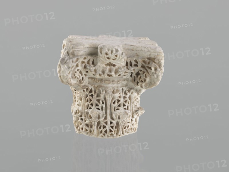 Column capital from Madinat az-Zahra, Islamic/Arabic art, Caliphate of Córdoba 929–1031, Al-Andalus, 10th century; architectural element—capital with acanthus ornament; height 22.3 cm, width 23.3 cm, depth 24.0 cm, weight 15.5 kg., An off-white carved stone fragment with intricate openwork patterns sits against a neutral gray background, showing a weathered top surface and a decorative band with leafy and floral motifs; the carving displays rounded forms, pierced voids, and textured surfaces, and the piece casts a faint reflection on the smooth gray surface below, with visible tones of off-white, light beige, and subtle gray.