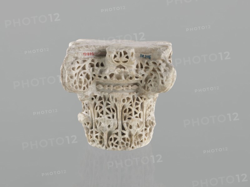 Column capital from Madinat az-Zahra, Islamic art, Arabic art, Arabic culture, Caliphate of Cordoba 929–1031, Al-Andalus, architectural element capital with acanthus ornament, 10th century, Madinat az-Zahra, Al-Andalus; height 22.3 cm, width 23.3 cm, depth 24.0 cm, weight 15.5 kg., A carved stone capital with intricate openwork patterns sits against a plain gray background, showing a weathered surface with varied beige and off-white tones and hints of light brown; the upper blocky section has rounded protrusions and floral-like perforations, while the lower cylindrical portion features vertical, vine-like carvings and leaf-shaped cutouts creating a lace-like appearance; subtle shadows and a faint reflection appear beneath the object, and overall the colors visible are gray, beige, off-white, light brown, and a trace of darker gray.