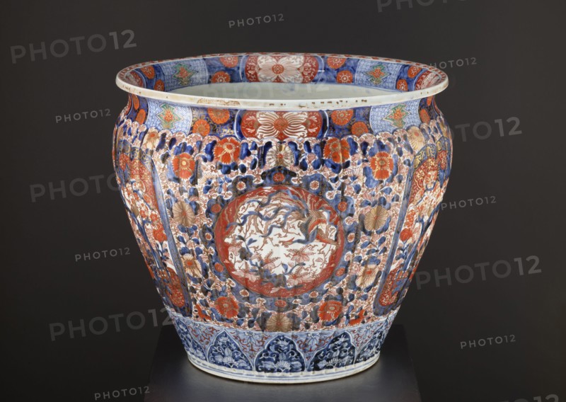 Flowerpot, ca. 1870–1880, Japan Arita. Porcelain jardiniere vessel with colored enamel and Imari palette; height 50.5 cm, diameter 58.0 cm, weight 28.0 kg. Decoration: floral ornaments including chrysanthemums, sakura, peonies, plant tendrils and geometric ornaments; kachôga bird-and-flower imagery featuring phoenixes/hoo and spotted pheasants. Style: Meiji 1868–1912; function: plant ornament., A large decorative ceramic vessel sits against a dark background, showing a bulbous shape with a flared rim and detailed painted ornament. The vessel displays a dense overall pattern of floral motifs and stylized foliage in shades of blue, red, and gold, with areas of white showing through the painted designs. A central roundel on the front contains a more delicate scene rendered in muted blue and red tones surrounded by a red border. Bands of patterned decoration encircle the rim and the base, featuring geometric and floral elements in blue and red with touches of green and gold. The surface al...