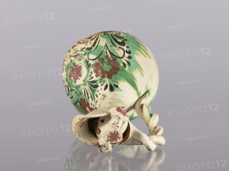 Jug, ceramic glazed jug with braided handle, filter, curved spout, neck cover with rosette; decorated with floral ornament; Ottoman Turkish, 19th century Çanakkale, Turkey; height 33.5 cm, diameter 15.7 cm., A ceramic object lies on its side against a neutral light gray background, showing an overall cream base color with areas of green and patches of reddish-brown and black decorative paint; the surface has floral and abstract painted motifs, and there is a twisted handle attached to the body with sculpted, rounded elements at the end, with some worn and chipped painted areas revealing the underlying cream color, and the object casts a faint reflection on the glossy surface beneath it.