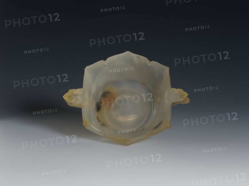 Bowl, 19th century, Qing 1644–1911, China. Spotted agate vessel with yellow-rusty stains and black dots. Hexagonal in the upper part, on a ring foot. Handles shaped as leafy branches with a flower. Morning glory motif. Height 4.7 cm, diameter 8.0 cm., A translucent, pale yellowish bowl with a smooth rounded interior and irregular petal-like edges sits centered against a dark gray to light gray gradient background; the bowl has a glossy surface with subtle milky swirls and warmer amber and honey-colored patches, and a small floral-shaped handle on each side that share the same translucent yellow and amber tones, casting a soft shadow on the lighter foreground.