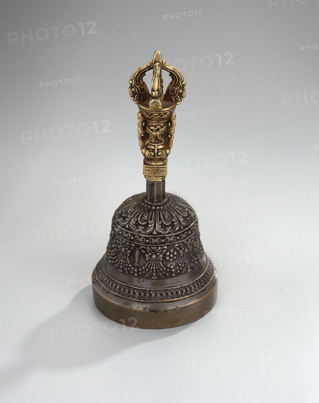 Bell;, Bowl with a lambrequin border with mascarons. Handle with a bust of a Bodhisattva carrying a vajra. Ceremonial object bell. Tibetan Buddhist / Himalayan art; vajras, bodhisattva iconography; heads; musical instruments — percussion idiophone. 18th–1st half of the 20th century; Tibet possibly. Height 14.9 cm; width 7.15 cm., A metal handbell stands on a pale gray background, with a dark bronze bell body decorated with ornate raised floral and patterned designs and a rounded base showing a lighter brownish-bronze rim; the handle is a contrasting golden color with sculpted intricate shapes and curved elements, and the bell casts a soft gray shadow to one side.
