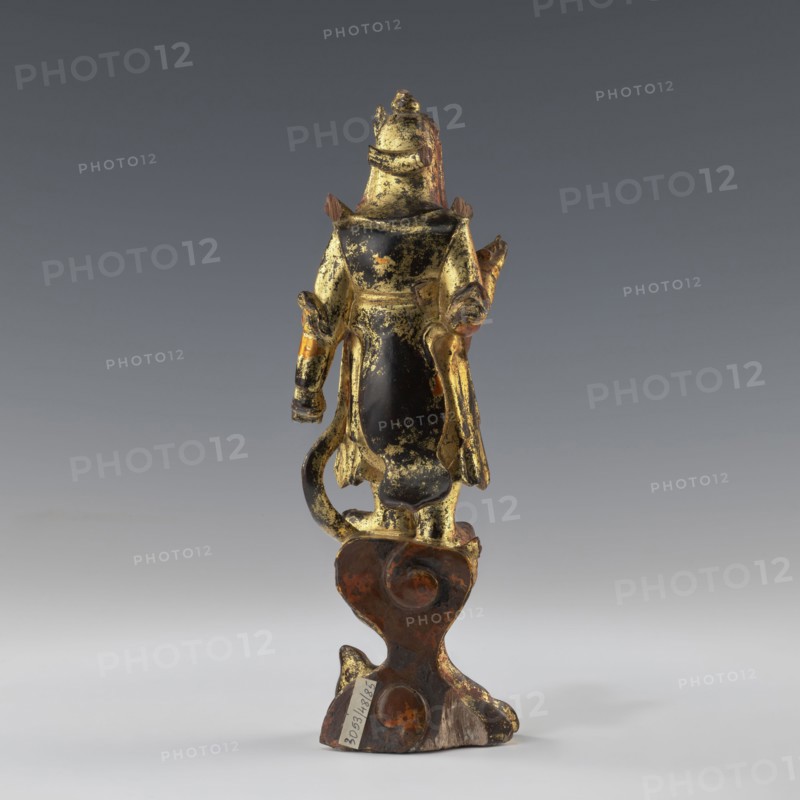 Guardian of Heaven, figurine, art of Southeast Asia, Vietnamese culture, 1st half of the 19th century, Vietnam, sculpture, height 22.8 cm, width 7.2 cm, depth 6.2 cm, A small standing figurine seen from the back, primarily showing a mix of gold and black surfaces with worn and flaked areas revealing brown and reddish tones; the figure wears a long garment with flowing panels and has articulated-looking arms and a tail-like curved element; it stands on an irregularly shaped base that is brown with patches of darker and lighter rust-colored areas and some exposed wood tone; the background is a smooth gradient of gray.