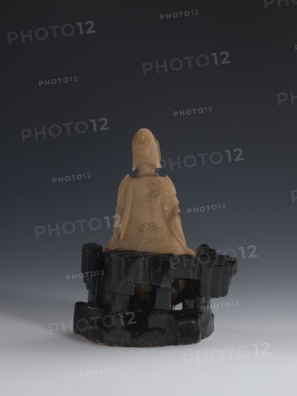 Guanyin Giving Birth to a Son on a Rock; sculptor unknown. Figurine of the goddess made of light coffee-colored alabaster, mounted on a stone base feldspar with red and pink spots, painted with black lacquer. Qing dynasty 18th/19th century, China. Chinese Buddhist subject: Goddess with a rosary in her right hand, holding a child with her left hand. Sculpture; height 18.2 cm, width 13.8 cm., A small carved figure seated on a carved base, viewed from behind, with a smooth light beige material for the figure and a glossy black material for the irregular, blocky base; the figure wears a draped garment with carved decorative patterns and a hood or headdress, and the base has an angular, stacked appearance with chipped edges and a reflective surface; the background is a smooth gradient from dark gray at the top to lighter gray at the bottom, and the visible colors are beige, black, gray, and a few subtle brownish tones on the base.