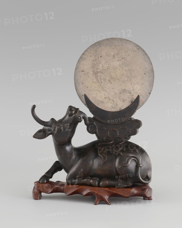 Mirror with TLV decoration on a pedestal in the form of a qilin on a base; Chinese, Qing dynasty 1644–1911. Mirror: cast from chiseled bronze, decoration in flat relief with a centrally placed knob enclosed in a square, four T-shaped motifs attached, and L- and V-shaped motifs at the edge. Pedestal: qilin cast from chiseled bronze; base: carved wood. 18th century mirror cast from an earlier product, possibly 6th century–?. Sculpture/mirror. Height 26 cm; diameter 16.2 cm., A sculpted reclining animal with curved horns rests on a carved wooden base, the animal appearing dark brown to black with a smooth, slightly polished surface and carved decorative lines on its body and tail; behind and above it is a large round disc that is mottled light beige to gray, held by a crescent-shaped support that is darker in color, and the background is a soft gradient of light gray.