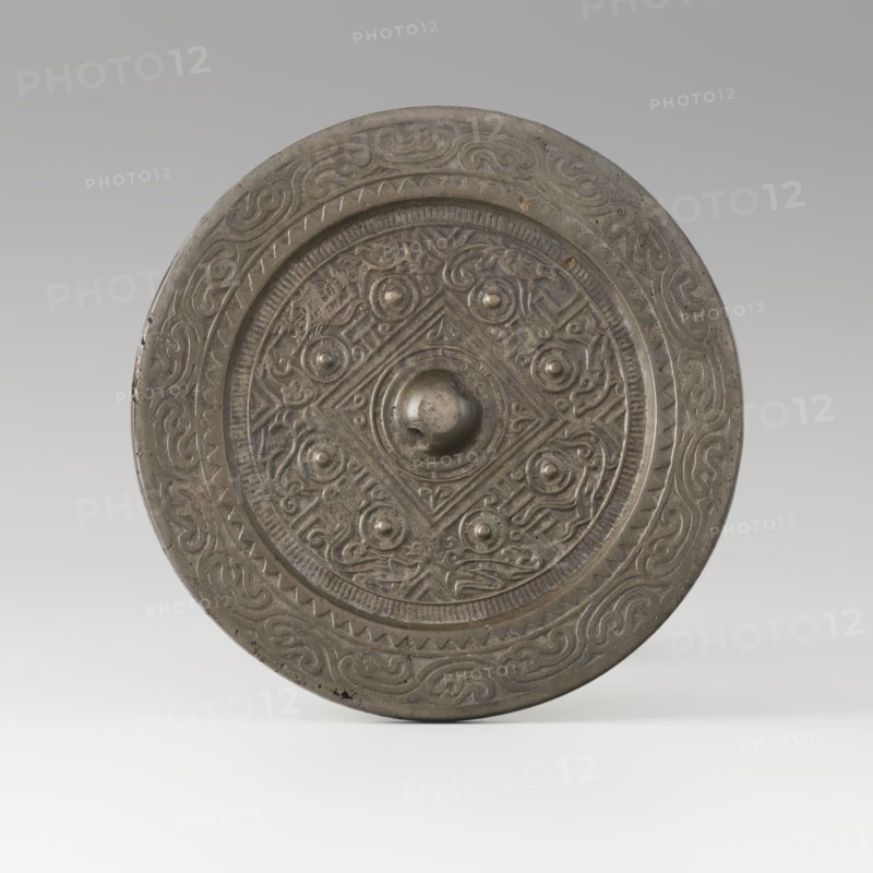 Mirror with TLV decoration on a pedestal in the form of a qilin on a base. Chinese, Qing dynasty 1644–1911. Materials: cast and chiseled bronze mirror with TLV flat-relief decoration—centrally placed knob enclosed in a square with four T-like motifs and L- and V-shaped motifs at the edge; unicorn qilin cast from chiseled bronze; carved wooden base. 18th century mirror cast from an earlier product, possibly 6th century–?. Sculpture/mirror. Height 26 cm; diameter 16.2 cm., A circular metal object with a raised central boss and concentric decorative bands, showing a patina of gray and dark gray tones with areas of brownish discoloration; the central area features a geometric motif with a diamond shape and a group of raised rounded knobs surrounded by intricate repeating scrollwork and curvilinear designs; the outer band contains a repeating wave-like pattern and a zigzag border, all rendered in muted metallic gray and darker shadowed gray, set against a pale gray gradient background.