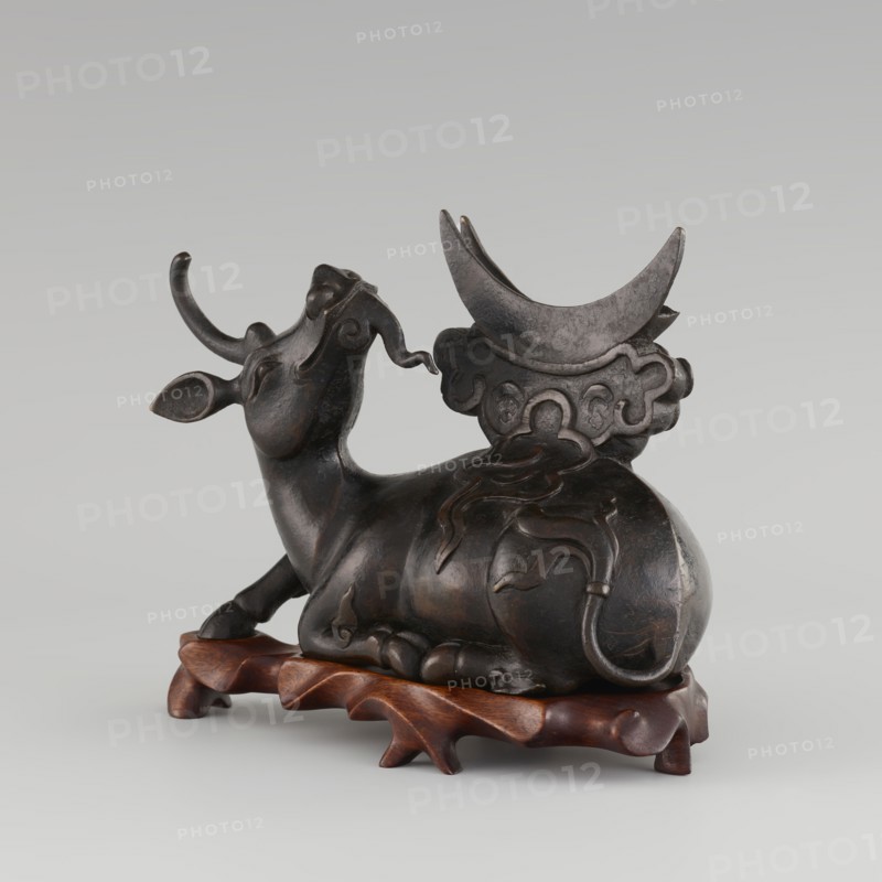 Mirror with TLV decoration on a pedestal in the form of a qilin on a base; Chinese, Qing dynasty 1644–1911; mirror cast from chiseled bronze with decoration in flat relief: centrally placed knob enclosed in a square with four T-shaped motifs attached, and L- and V-shaped motifs at the edge; unicorn qilin cast from chiseled bronze; carved wooden base; 18th century mirror cast from an earlier product, possibly 6th century–?; sculpture, mirror; height 26 cm, diameter 16.2 cm; bronzes, mirrors, castings, qilin iconography., A dark brown sculptural figure of a reclining bovine with curved horns and a raised head sits on a carved reddish-brown wooden base; the bovine has smooth rounded forms, stylized flowing decorative lines along its body and tail, and a large crescent-shaped element rising behind it that rests on a cloud-like carved support, all rendered in shades of dark brown and black with subtle highlights.
