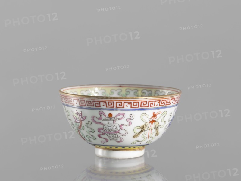 Bowl, gift, Qing dynasty 1644–1911, 19th century, China. Colored porcelain bowl on a foot, height 3.6 cm, diameter 6.6 cm. Decorated on the outside with a meander stripe and Buddhist symbols cranes, endless knot, lambrequin pattern; Chinese ceramics, bowls; related to Boxer Rebellion 1899–1901., A small porcelain bowl with a glossy white surface sits against a plain gray background, decorated around its exterior with a repeating band of geometric motifs in red near the rim, a thin gold trim at the top edge, and a narrow yellow band with blue dots near the foot; the bowl shows painted ornamental designs in soft green, pale pink, lavender, orange, and blue including stylized floral and ribbon-like shapes and a small rendered vessel, with delicate gold accents and a reflective sheen on its curved surface.