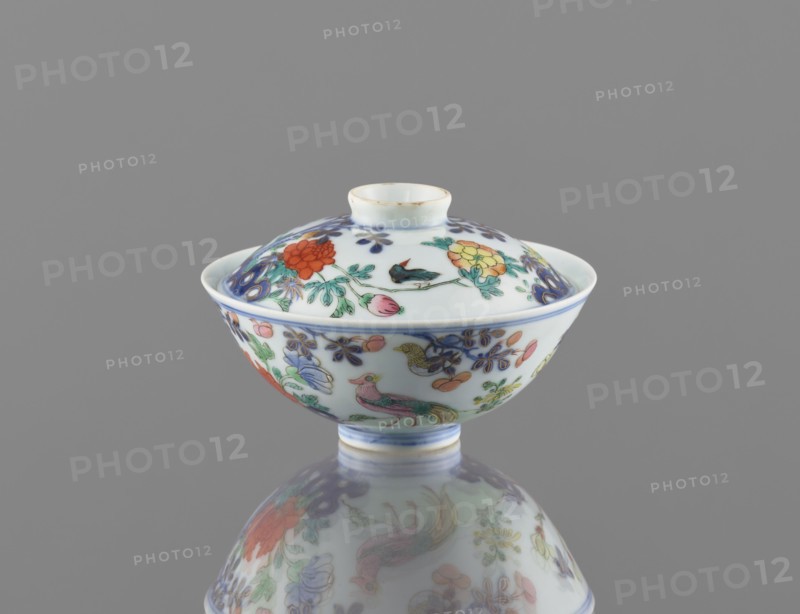 Bowl with a lid, Chinese, Qing dynasty, Qianlong period 1736–1795. Colored porcelain vessel decorated in a garden setting with boulders, trees, peonies, magnolias, chrysanthemums; two pheasants and a butterfly. Signed. Height 7.5 cm; diameter 11.7 cm; weight 0.2 kg., A lidded porcelain bowl with a rounded foot sits on a reflective surface, showing a mirrored image beneath; the bowl and lid are white with painted floral and bird motifs in colors including red, yellow, green, blue, purple, pink, and orange, and there are areas of gold or brownish rim detailing; the background is a uniform gray.