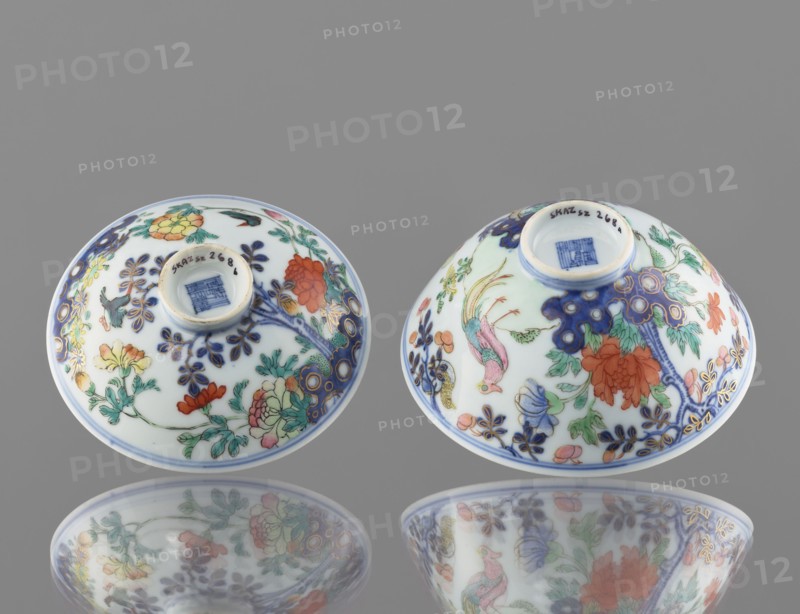 Bowl with a lid, Chinese; Qing dynasty, Qianlong period 1736–1795; colored porcelain; decoration: in a garden setting boulders, trees, peonies, magnolias, chrysanthemums two pheasants and a butterfly; signed; vessel; height 7.5 cm, diameter 11.7 cm, weight 0.2 kg., Two small porcelain bowls are shown upside down on a smooth reflective surface, each decorated with floral and bird motifs. The bowls are primarily white with intricate painted designs in a palette that includes blue, green, red, orange, pink, yellow, and gold accents. One bowl features a prominent red-orange flower and leafy green stems with blue ornamental detailing, while the other shows a colorful bird with pink and green plumage among similar floral patterns and blue branch-like decorations. Both bowls have a ringed foot and a central recessed base area, and their painted surfaces and the gray background are reflected beneath them.