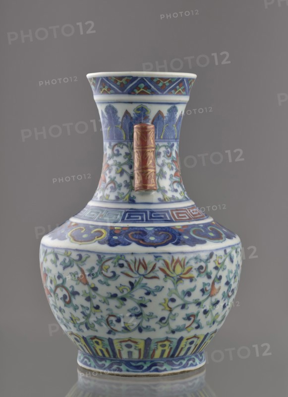 Vase with threads in the shape of bamboo twigs; maker, Chinese, Qing dynasty, Qianlong period 1736–1795; colored porcelain; decoration: lotus tendril on the belly, artemisia leaves, lambrequin ornament on the shoulders, geometric and plant borders on the neck; plant motifs; vessel; height 20 cm, diameter 14.5 cm, weight 0.8 kg., A decorative ceramic vase with a flared neck and bulbous body sits against a gray background, featuring a palette of blue, white, green, yellow, red, and gold; the neck shows stylized leaf motifs in blue and green and a vertical cylindrical red and gold accent attached to its front, while the shoulder and body display scrolling floral vines with lotus-like flowers in red, yellow, and green on a white ground, bordered by geometric and cloud-like bands in blue and red near the top and a band of vertical panel motifs in blue, yellow, and red near the base, with a narrow blue patterned rim at the foot.
