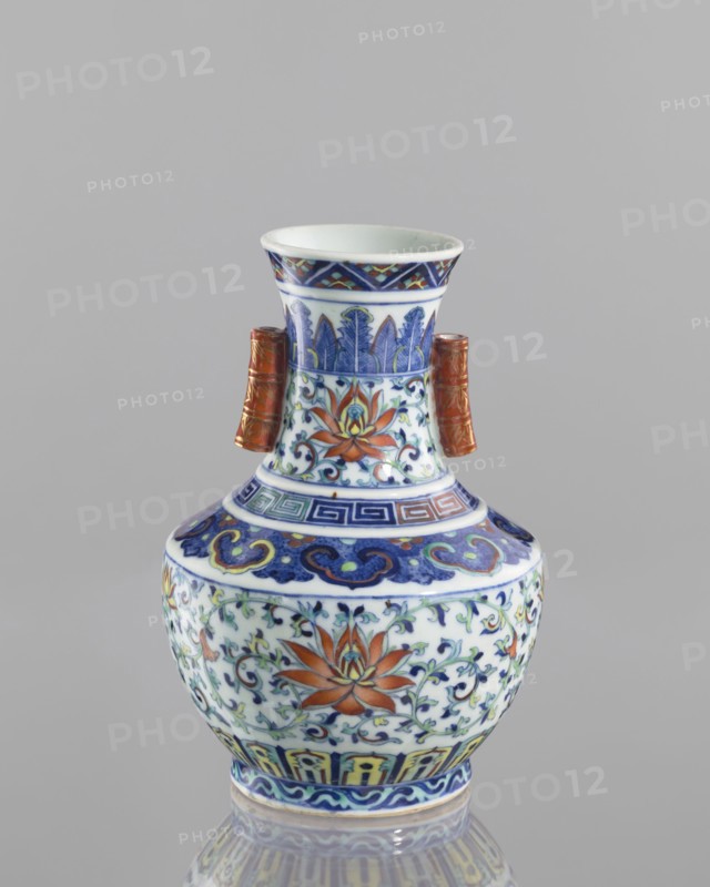 Vase with threads in the shape of bamboo twigs; colored porcelain; Chinese; Qing dynasty, Qianlong period 1736–1795; decoration: lotus tendril and artemisia leaves on the belly; lambrequin ornament on the shoulders; geometric and plant borders on the neck; motifs: bamboos, lotuses, lotus tendril ornament, meander ornament, geometric ornaments; vessel; height 20.0 cm, diameter 14.5 cm; maker, function: vase; subject: flowers., A ceramic vase sits against a plain gray background, featuring a bulbous body, a narrowed neck, and a flared rim, with a pair of short tubular handles on the neck. The surface is decorated with ornate painted floral and foliate motifs in colors including blue, white, orange, green, yellow, and a reddish-brown on the handles, with bands of geometric and curving patterns encircling the neck and shoulder. The primary floral motifs show stylized flowers with orange petals and green leaves amid scrolling blue vines, and the rim and base areas display repeating decorati...
