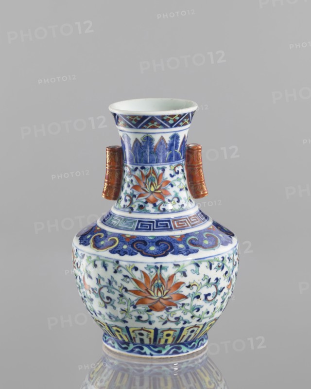 Vase with threads in the shape of bamboo twigs;, colored porcelain; Chinese, Qing dynasty, Qianlong period 1736–1795; decoration: lotus tendril and artemisia leaves on the belly; lambrequin ornament on the shoulders; geometric and plant borders on the neck; motifs: bamboos, lotuses, lotus tendril ornament, meander ornament, geometric ornaments; function: vessel vase; height 20.0 cm, diameter 14.5 cm; inscriptions/notes: flowers., A small decorative vase sits against a neutral gray background, showing a bulbous body with a narrower neck and flared rim, decorated with intricate painted floral and geometric patterns in colors including blue, white, orange, green, yellow, and brown, and featuring two short cylindrical side handles in a brownish-gold tone; the surface displays stylized lotus-like flowers and scrolling vine motifs, bands of geometric fret and cloud-like patterns, and a glossy finish that creates a subtle reflection beneath the vase.