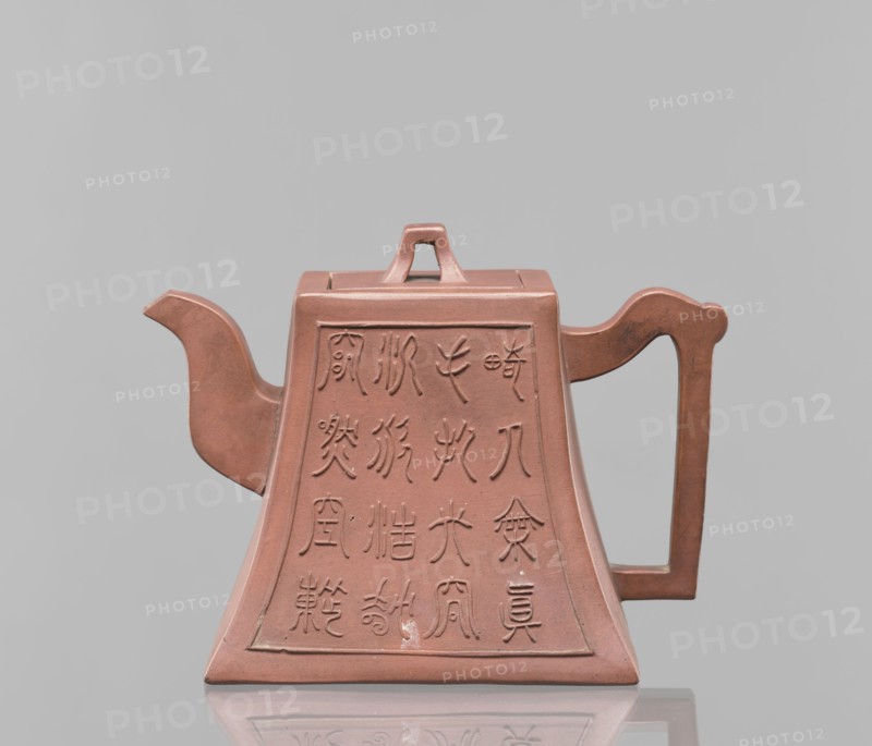 Teapot with a lid, tea, Harbin China, ceramics, Chinese art, Chinese culture, Qing 1644–1911, Yixing style, inscriptions, red-brown stoneware, 19th century, China, vessel; height 12.2 cm, width 16.3 cm, A reddish-brown teapot with a trapezoidal body and a flat top, featuring a short triangular lid handle, a curved spout on the left, and a rectangular handle on the right; the front and sides show recessed panels with incised decorative markings, and the object sits on a reflective surface against a pale gray background with visible colors including reddish-brown and gray.