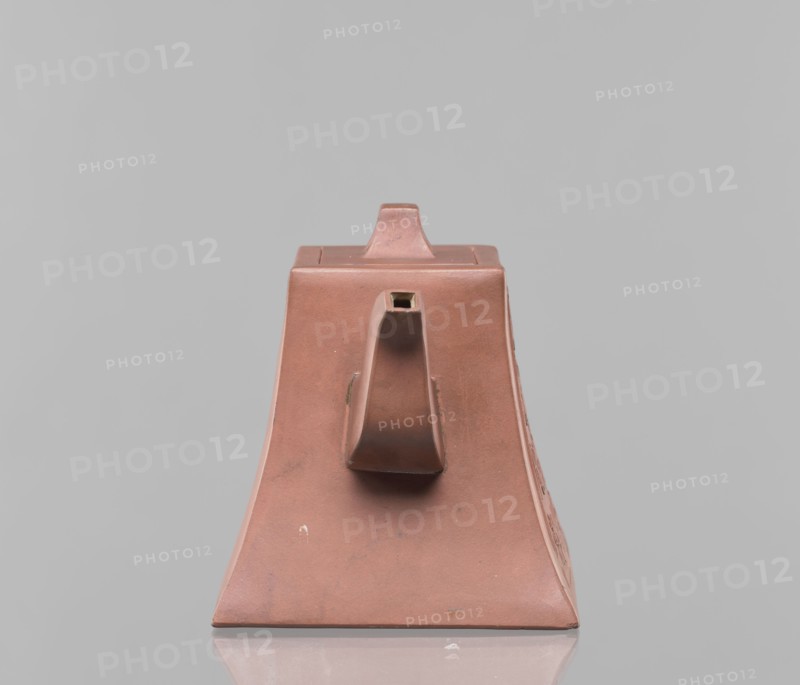Teapot with a lid, teapot for tea, Harbin China, ceramics, Chinese art, Chinese culture, Qing dynasty 1644–1911, Yixing style, red-brown stoneware, 19th century, China, vessel, height 12.2 cm, width 16.3 cm, inscriptions., A trapezoidal ceramic object in a muted reddish-brown color sits centered on a reflective surface against a pale gray background. The object has a flat top with a small raised rectangular knob, and a central protruding spout-like feature on its front face that is hollow at the tip. The sides slope outward toward the base, and the surface shows subtle variations in tone and a few small blemishes and scuffs in darker and lighter reddish-brown shades. The reflection of the object's base appears faintly on the surface below, and overall the visible colors are reddish-brown and gray with minor darker and lighter reddish-brown marks.