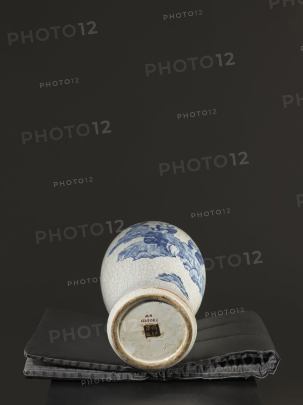 Vase, figurative scenes, ceramics, gardens, Chinese art, Chinese culture, blue-and-white porcelain, games, cracklework decoration, play, Qing 1644–1911, 18th–19th century Jiaqing period, 1797–1820, Qing dynasty, China, vessel, height 54.5 cm, width 9.8 cm, A ceramic vase lying on its side on a folded fabric pad, the vase showing a crackled white glaze with blue painted motifs and a rounded base with an unglazed foot rim, the fabric pad appearing dark gray and black with stitched seams, all set against a solid dark background with shades of black and deep gray.