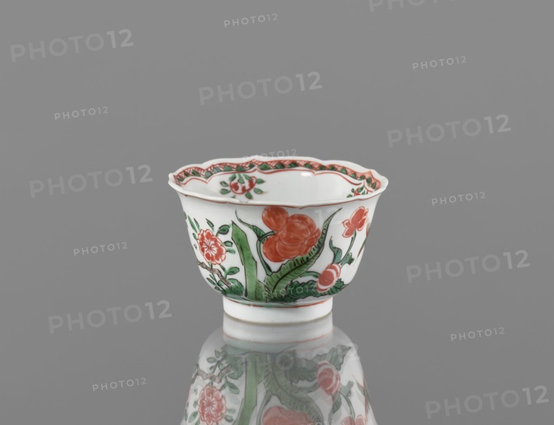 Bowl, porcelain with colorful overglaze enamels in Kangxi style Qing dynasty, 1662–1722. Decorated with floral motifs, plum trees, and bird images. Vessel, height 5.4 cm, diameter 8.8 cm; China., A small white porcelain cup with a scalloped rim sits on a reflective surface against a gray background; the cup is decorated with floral motifs in red and green, including a few large red flowers and several green leaves, and a patterned red-and-green border runs just inside the rim, with the cup’s reflection visible beneath it.