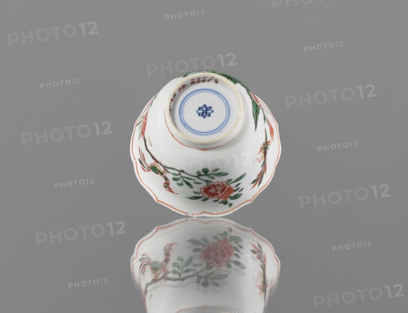 Bowl, ceramic vessel, colorful porcelain in Kangxi style Kangxi period, 1662–1722, Qing dynasty 1644–1911. Decorated with floral motifs, plum trees and bird images. China; height 5.4 cm, diameter 8.8 cm., A white porcelain bowl with scalloped edges sits upside down on a reflective gray surface, showing painted floral and bird motifs in red, green, and black with blue circular detail at the base and a reflection of the bowl visible below; the background is gray.