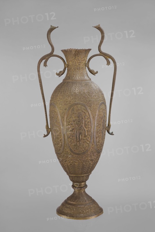 Vase, repossession; Vishnu; Indian art; Indian culture; cobras snakes; plant ornament; geometric ornament; figural scenes; fish; halos; maces; religion; snakes; processions; flutes; clubs. On the belly six ovoid reserves with figural decoration four female figures with wreaths, two images of Vishnu; plant and animal motifs on the entire surface. 19th century, India. Vessel. Height 65.0 cm; width 27.5 cm; weight 2.95 kg., The image shows an ornate metal vase with a tall, elongated body and a flared neck, finished in shades of gold and bronze; the surface is covered in detailed raised decoration including repeated oval panels with human figures and elaborate floral and foliate patterns; a pair of long, curved handles rise from the shoulders and loop upward with stylized animal head terminals, matching the gold-bronze coloration; the vase stands on a pedestal base that echoes the decorative motifs and colors; the background is a smooth, neutral gray.