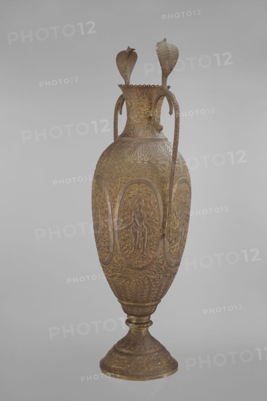 Vase, 19th century, India; vessel, height 65.0 cm, width 27.5 cm, weight 2.95 kg. On the belly six ovoid reserves with figural decoration: four female figures with wreaths and two images of Vishnu. Surface decorated overall with plant and animal motifs, cobras snakes, fish, geometric and plant ornament, figural scenes, halos, maces, processions, flutes, clubs; religion Vishnu., An ornate tall vase with a rounded body and a flared neck resting on a stepped pedestal base, rendered in shades of brown and muted gold; the surface is richly embossed with repeating floral and foliate patterns and several oval medallions each containing a raised human figure in relief; two long, curved handles extend from the neck to the shoulder and terminate near the body, and two decorative leaf-shaped finials rise above the rim; the background is a neutral gray.