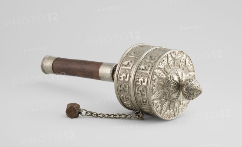 Prayer wheel, Tibetan Buddhist art, Himalayan culture, Himalayan art, scrolls, Buddhist art, 19th century, Tibet, cult object, height 26.5 cm, An ornate handheld object lies on a neutral background, showing a cylindrical carved metal head with raised floral and geometric motifs and a rounded, petal-like protrusion; the head is silvery in color with darker shadowed recesses. Attached to the head is a short metal chain leading to a small faceted weight that is brownish in color. Extending from the head is a wooden handle that is brown with visible grain and a slightly darker patina, capped at the end with a silvery metal band. The overall scene contains colors of silver, brown, and subtle gray tones in the background.