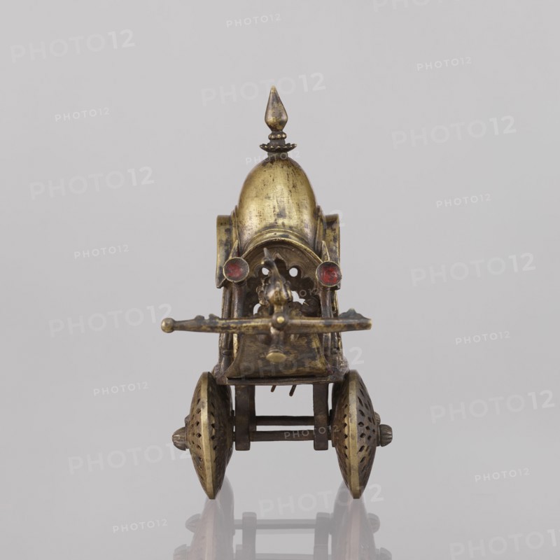 Miniature of a chariot;, 19th century, India; model; height 24.0 cm, width 28.0 cm. Temple chariot motif with peacocks and birds. A bird sits on a bow-shaped shaft. Under a canopy topped with a pike sit figures: Krishna feeding on butter; in front, in the driver's seat, there is also Krishna. Indian art; South Asian., A small metallic wheeled object viewed from the front, with a domed top and a pointed finial, showing a worn brass or gold-toned surface with darker patina; a pair of round red accents sit near the front of the dome; a horizontal bar protrudes across the front above an open lattice-like opening with curved decorative cutouts; two ornate spoked wheels with perforated patterns flank the lower sides, connected by a rectangular axle structure in a darker metal; the background is a smooth, light gray surface and the object casts a faint reflection below.