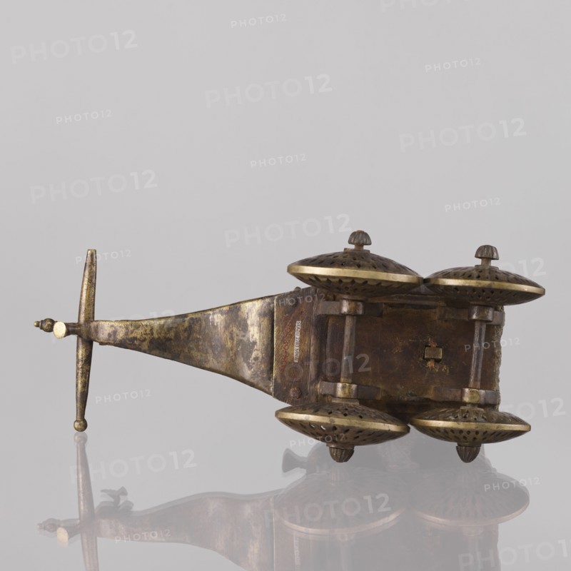 Miniature of a chariot; maker, 19th century India; model of a temple chariot depicting peacocks and birds. A bird sits on a bow-shaped shaft. Under a canopy topped with a pike sit figures: Krishna feeding on butter; in front, in the driver's seat, there is also Krishna. Materials/technique not specified. Height 24.0 cm, width 28.0 cm., An aged metallic object with a long tapered handle and a crossbar end lies on a reflective surface; the metal shows a mottled patina of brown, dark brown, and muted gold tones. Attached to the wider end of the object is a rectangular block from which a group of dome-shaped caps project; the domes are bronze-colored with darker perforated patterns and lighter golden rims, and each dome has a small rounded finial on top. The rectangular block and connecting posts share the same brown and gold worn metallic finish, and the reflection beneath the object repeats the brown, gold, and darker shadowed hues. The background is a smooth pale gray.