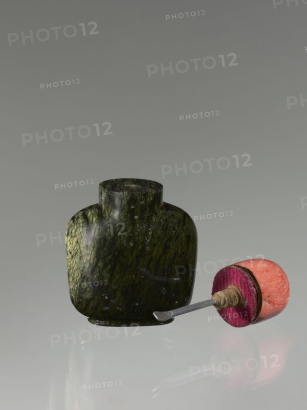 Snuffbox with spoon, Chinese, Qing dynasty 1644–1911, 19th century. Moon bottle form carved from rhodochrosite plug stone. Vessel; height 7.1 cm, width 4.6 cm, depth 2.6 cm., A smooth, dark green stone container with mottled lighter green veins sits on a reflective surface, accompanied by a small scoop tool that has a metallic shaft and a rounded end wrapped in twine attached to a cylindrical handle showing bright pink and salmon orange tones.