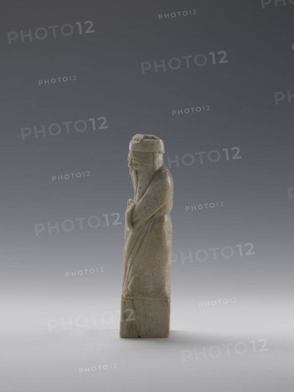 Sage, figurine; men; Chinese art; Chinese culture; Qing 1644–1911, 19th century; China; sculpture; height 16.8 cm, width 4.6 cm, depth 4.3 cm., A carved stone figure standing upright on a rectangular base, rendered in pale greenish-beige with faint areas of brownish veining and slight reddish stains, depicted in a long robe with flowing folds and a long beard, wearing a rounded hat and holding its hands together at the front, shown in profile against a smooth gradient background that transitions from dark gray at the top to light gray at the bottom.