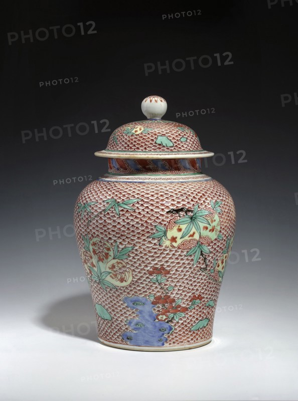 Vase with cover, 18th century, China; porcelain vessel with domed cover; Qing dynasty references to Ming/Wanli styles; ceramics decorated with fish scale ornament and pomegranates; backdated signatures; height 30.5 cm, diameter 19.5 cm; signed., A lidded ceramic jar with a rounded body and finial-topped lid stands on a light surface against a dark background. The jar is covered in a repeating scale-like pattern in red with areas of pale cream showing through, and it features painted floral and botanical motifs in green and pale yellow with touches of red. There are accents of blue and violet near the lower body and around the shoulder, and the lid has similar red scale patterning with green leaf-like shapes and a cream-colored knob with small red markings. The overall colors visible include red, cream, green, blue, pale yellow, violet, and black outlines.