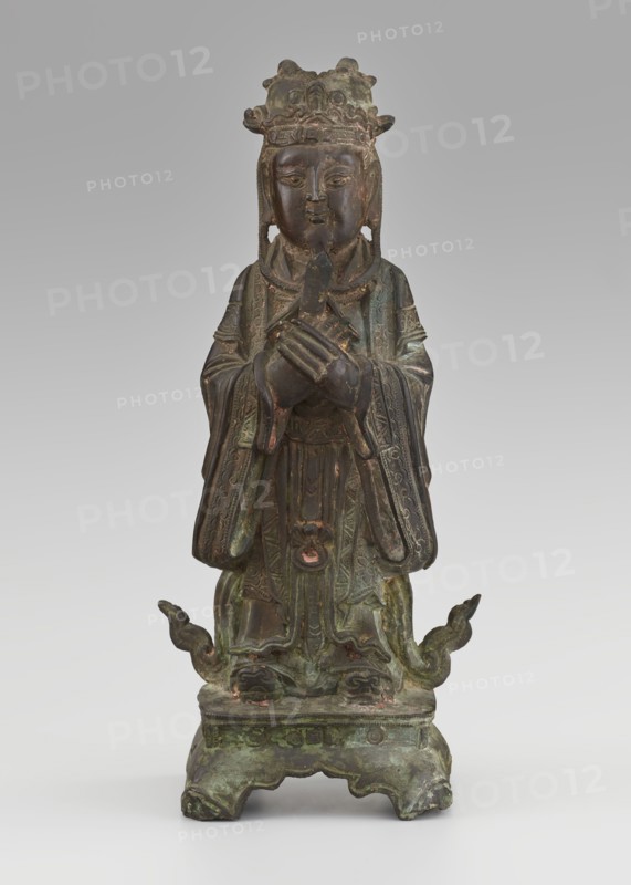 Dignitary — official; maker, figurine of a man; Chinese art; culture: Chinese; subject: scholars, sages, government officials; material: sculpture; date: 19th century Qing dynasty, 1644–1911; dimensions: height 28.5 cm, width 12.4 cm, depth 8.7 cm., A standing weathered statue with a patina of green, brown, and dark bronze tones depicts a robed figure wearing an ornate crown, holding a small object with both hands crossed at chest level; the face is serene with closed lips and finely carved facial features, and long earflaps or hair hang beside the face. The flowing robes display detailed carved patterns and layered sleeves with traces of darker brown and coppery highlights, and the figure stands on a square base that shows carved decorative elements and areas of green corrosion. Two upward-curving decorative elements extend from the base near the figure’s feet, and overall the surface shows aged texture, spots of verdigris green, dark brown, and hints of reddish-brown.