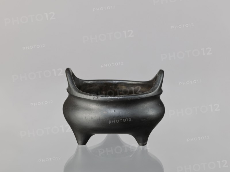 Li-type incense burner, cicadas and lion iconography, dragons, Chinese art/culture, Ming dynasty 1368–1644, Xuande period 1426–1435, ceremonial/ritual vessel, China, vessel, height 8.1 cm, diameter 11.1 cm, A small rounded vessel with a dark, almost black surface sits centered on a smooth, reflective surface; it has a wide, open top with two upward-curving rim extensions on opposite sides, a bulging body that narrows toward the base, and three short, tapered legs; the interior appears darker and slightly worn, and the surrounding background is a soft neutral gray.